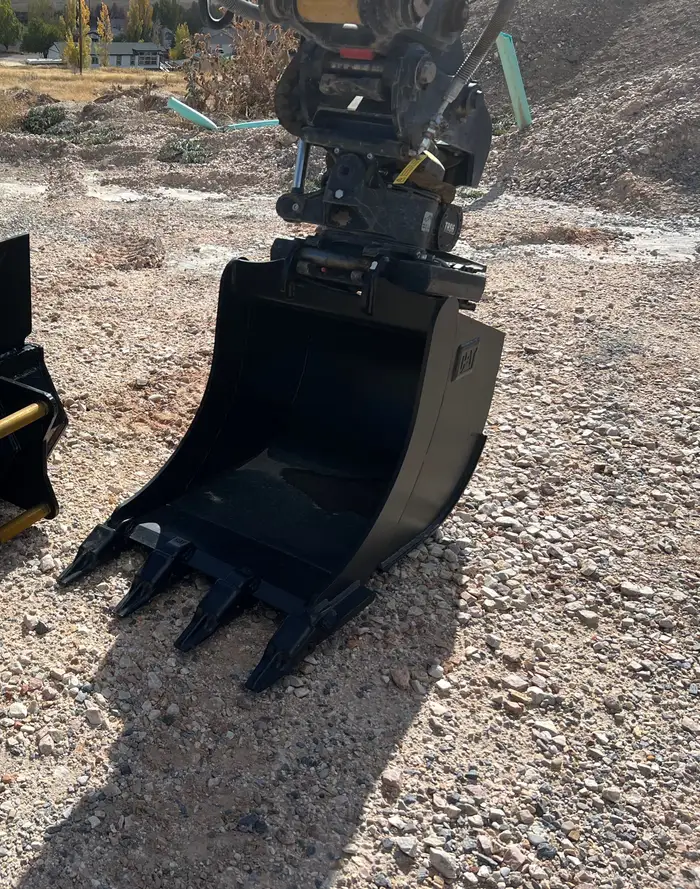 A digging bucket on a quick coupler resting on gravel, ready to swap onto an excavator