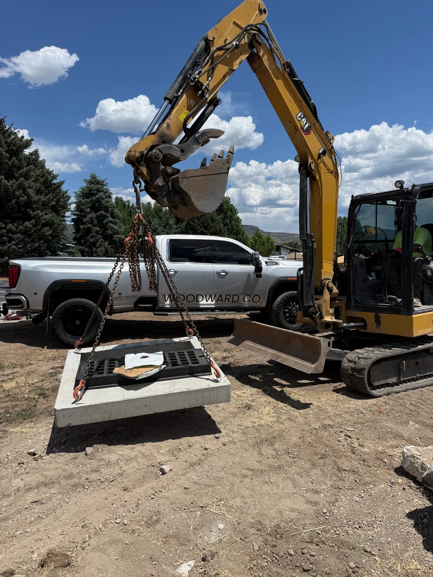 A mini excavator lifting a precast catch basin on chains