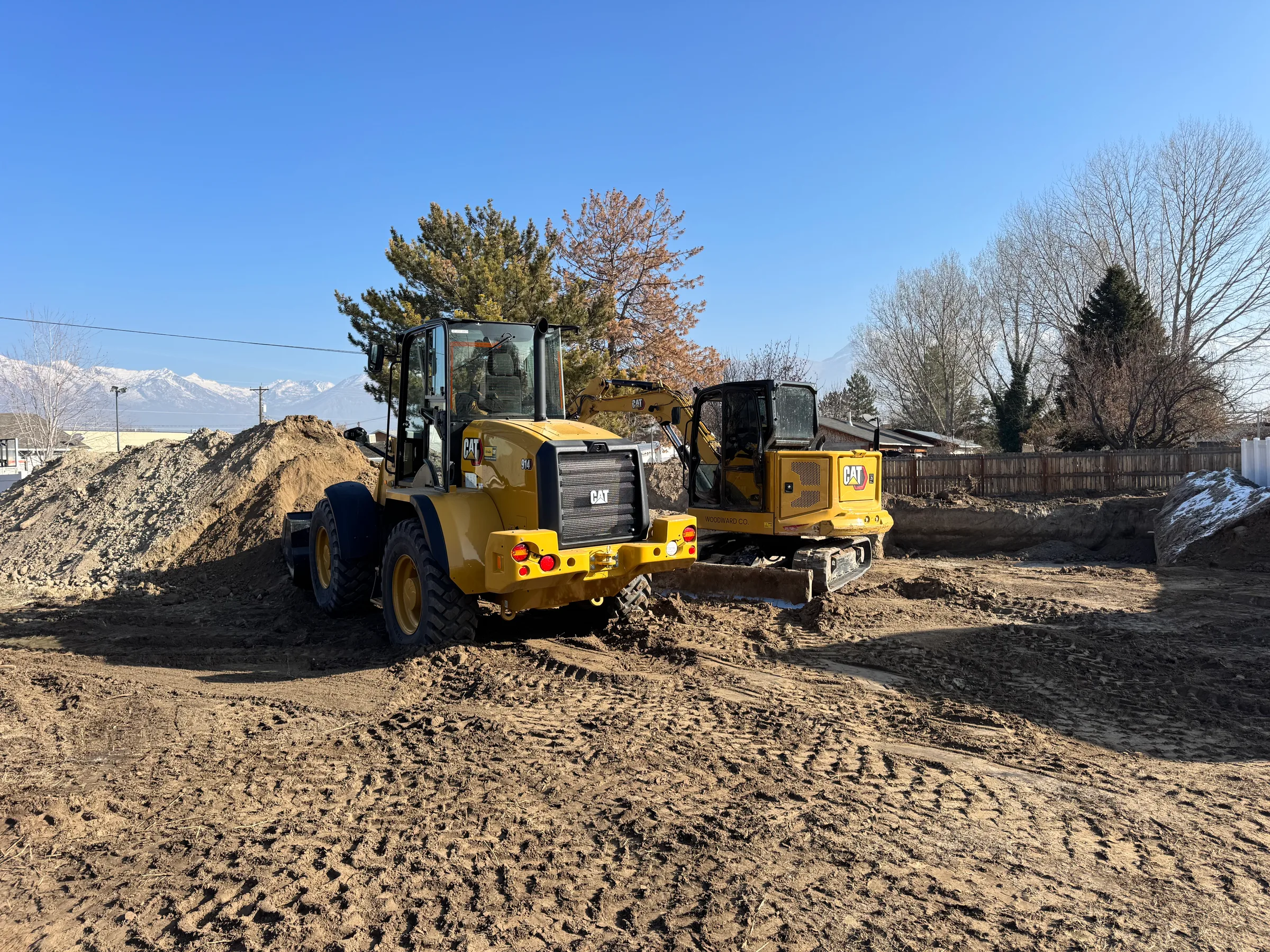 A wheel loader and excavator on a residential lot dig under snowy peaks