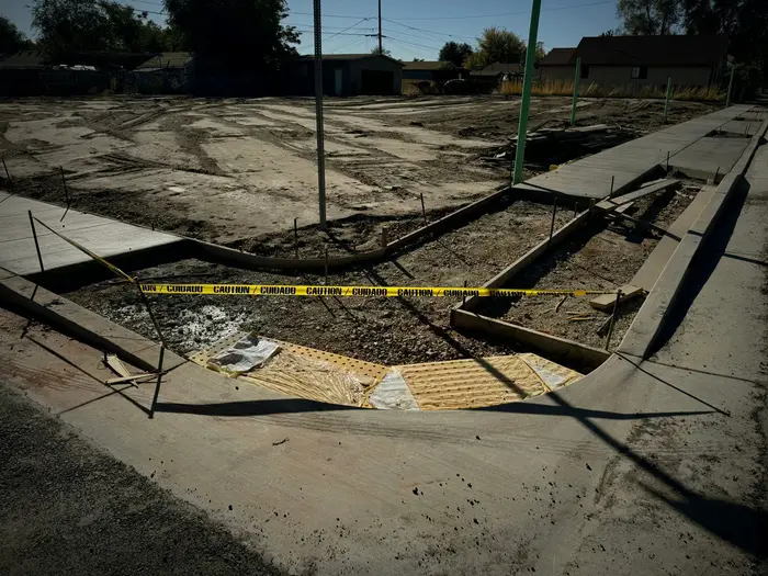 New concrete curb, gutter and sidewalk with an ADA ramp at the corner, caution tape across it and a graded lot behind