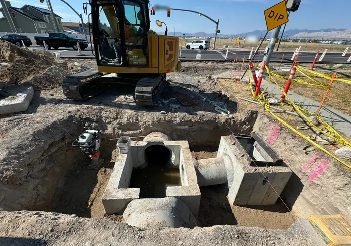 A precast concrete storm drain box with pipe connections in an open excavation at an intersection, a compactor and a mini excavator beside it