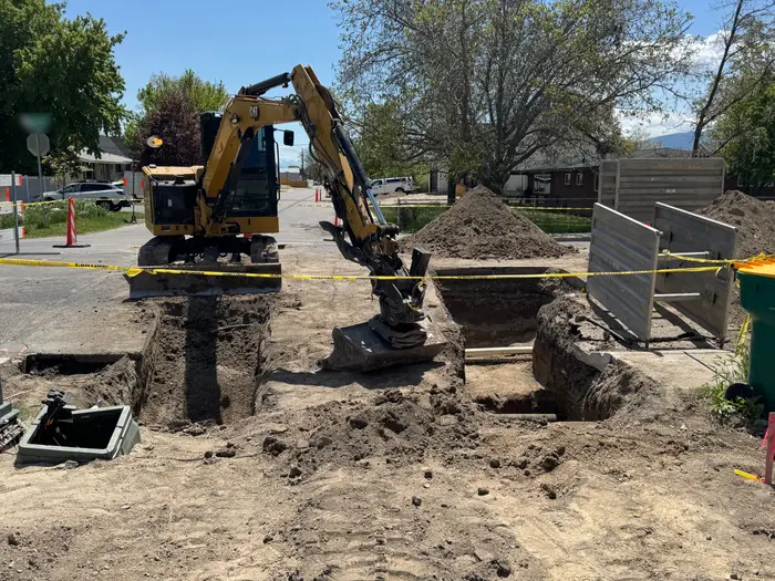 A mini excavator at a street corner beside two open trenches, aluminum trench shoring and caution tape