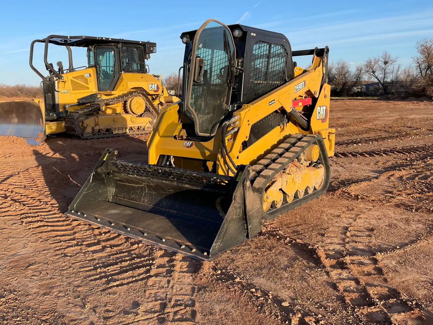 A yellow CAT compact track loader and a CAT dozer parked on freshly cleared red dirt in golden evening light