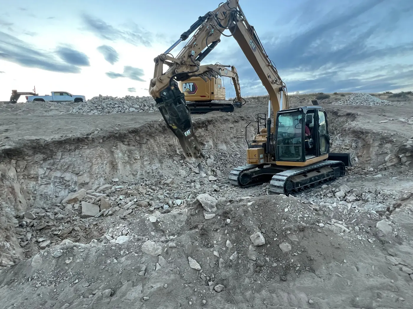 A yellow CAT excavator with a hydraulic breaker hammering rock in a basement pit at dusk, a second excavator behind