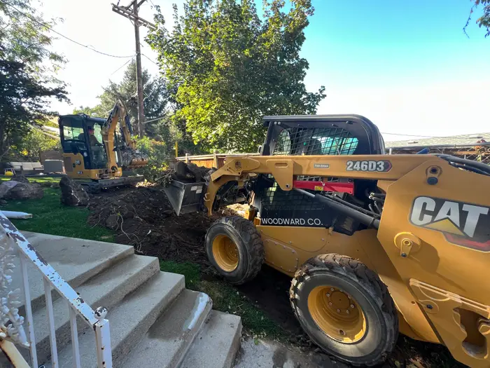 A yellow CAT skid steer carrying a bucket of roots and stumps across a front yard, a mini excavator digging behind