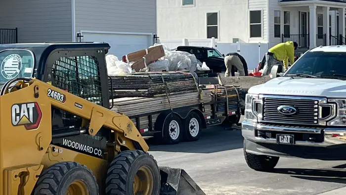 A yellow CAT skid steer and a white pickup on a new street while the crew unloads a trailer of lumber