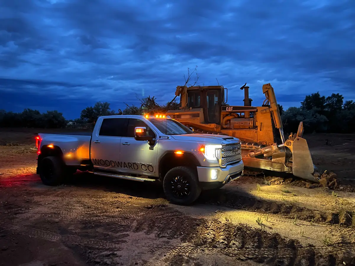 A silver crew pickup with its amber lights on beside an older CAT dozer at blue hour, dark clouds overhead