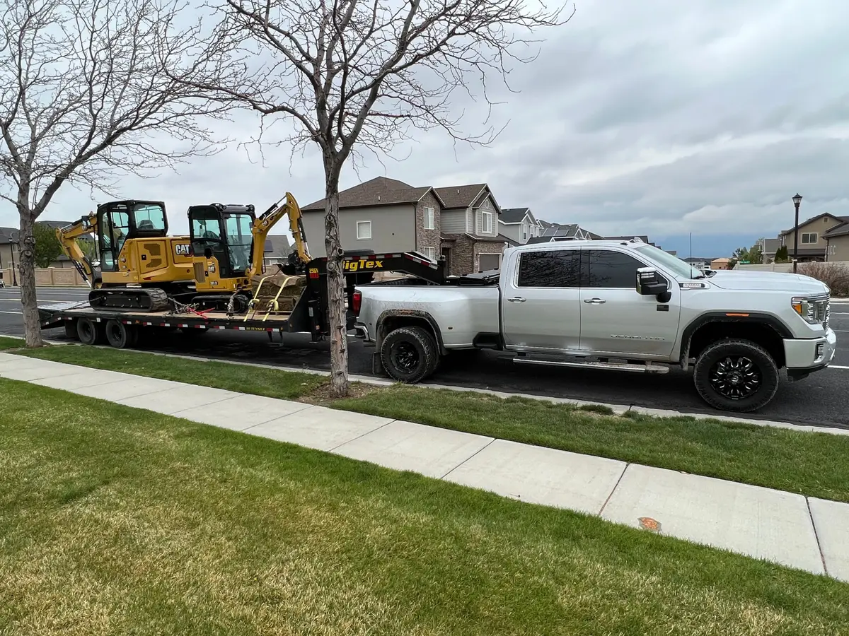 A silver dually pickup towing a gooseneck trailer with two yellow CAT mini excavators strapped down, parked on a residential street
