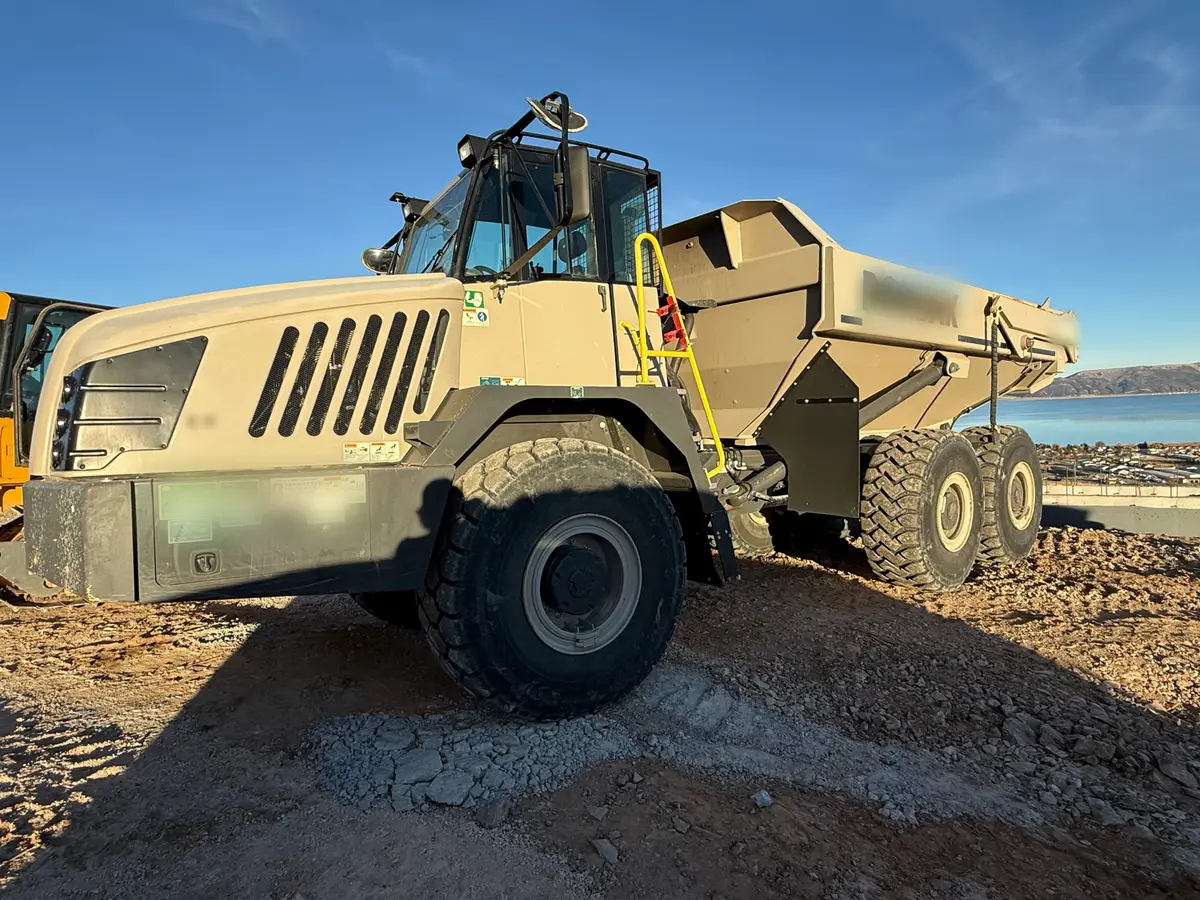 A tan articulated haul truck parked on a rocky hilltop job, a blue lake and hills behind it