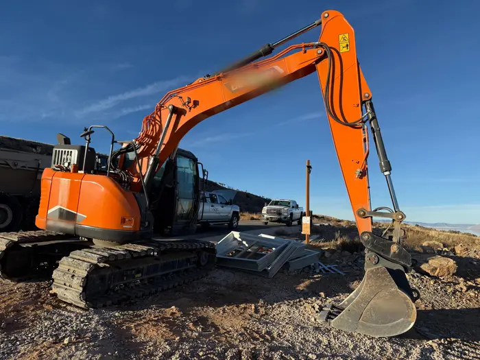 A rented orange excavator with its boom raised, stacked wall forms and two pickups behind it on a hilltop