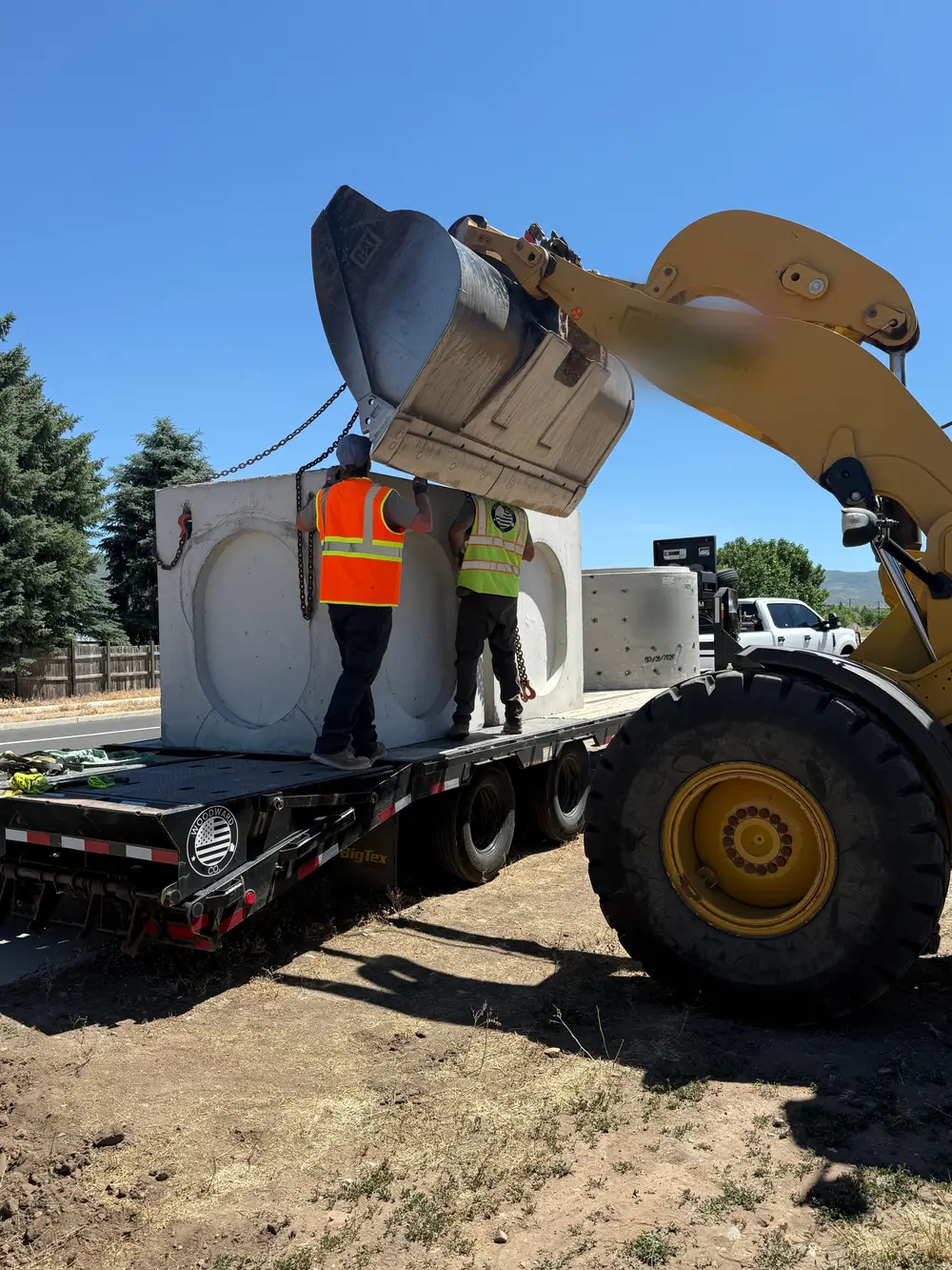 Two crew members in hi-vis vests guide a large precast concrete box on chains as a wheel loader lifts it off a trailer