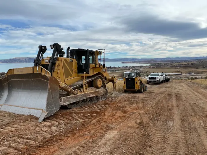 A big CAT dozer, a track loader and three white crew pickups lined up on a dirt road above a lake