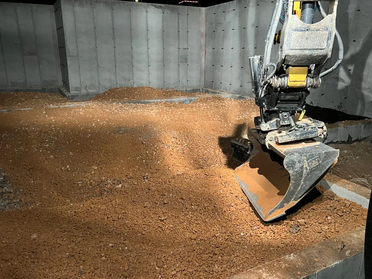 At night under work lights, an excavator bucket spreading gravel base inside a new concrete foundation