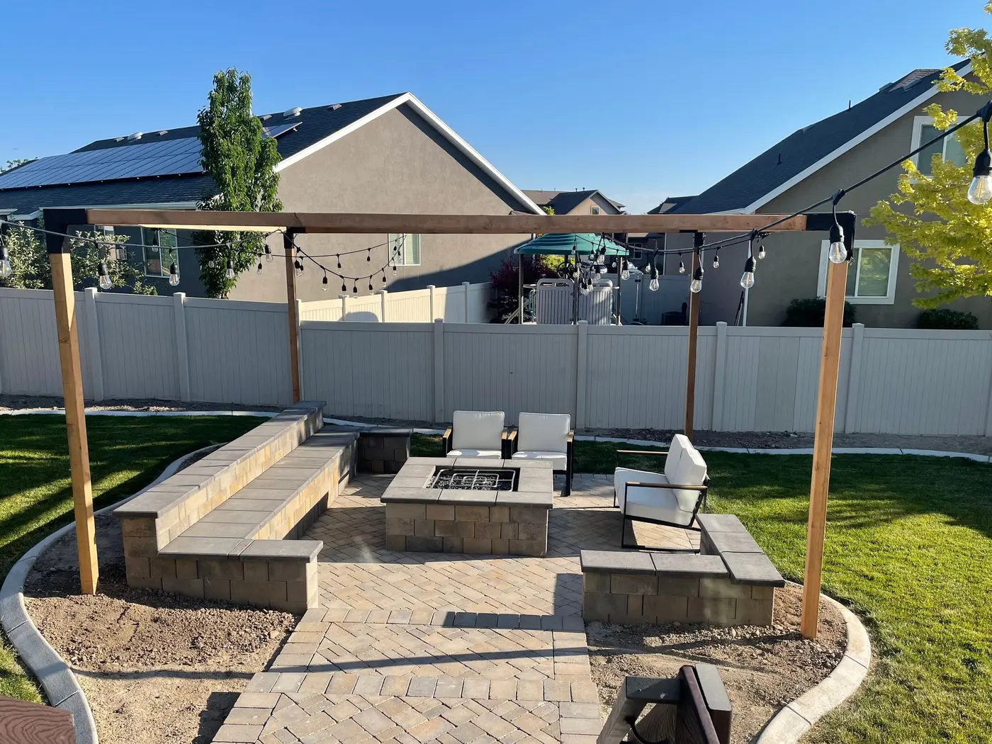The finished patio: chairs around the fire pit, string lights on cedar posts, curved curbing and green lawn