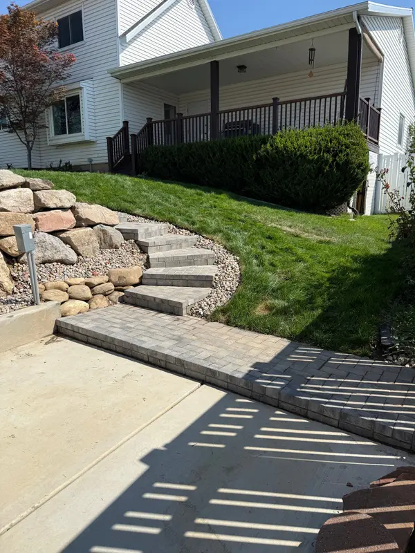 Curved paver steps climbing through a boulder retaining wall to a lawn and a covered porch