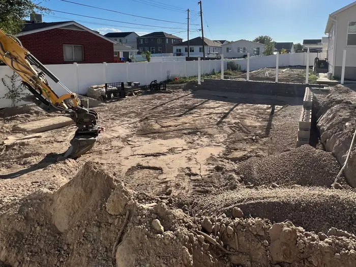 A new-home backyard dug out for retaining walls: a mini excavator arm on the left, a gravel-filled footing trench with the first blocks on the right, a white vinyl fence behind