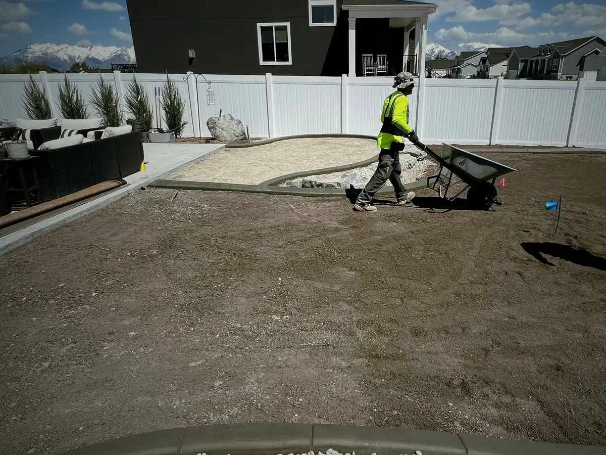 A crew member in a bright safety shirt pushes a wheelbarrow across a freshly graded topsoil backyard with new curbing, a white fence and snowy mountains behind