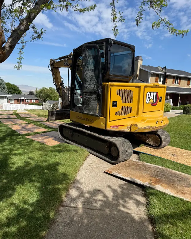 A yellow CAT mini excavator on a sidewalk with plywood sheets laid across the green lawn on both sides to protect the grass