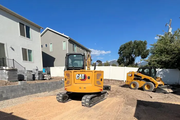 The same backyard a week later: a gray block retaining wall along the house, pallets of block by the steps, a yellow CAT mini excavator and skid steer on the graded yard