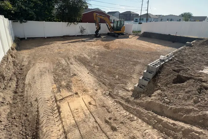 The same backyard two days later: graded dirt with tire tracks, the mini excavator at the back fence and a row of cinder blocks going in along the right side