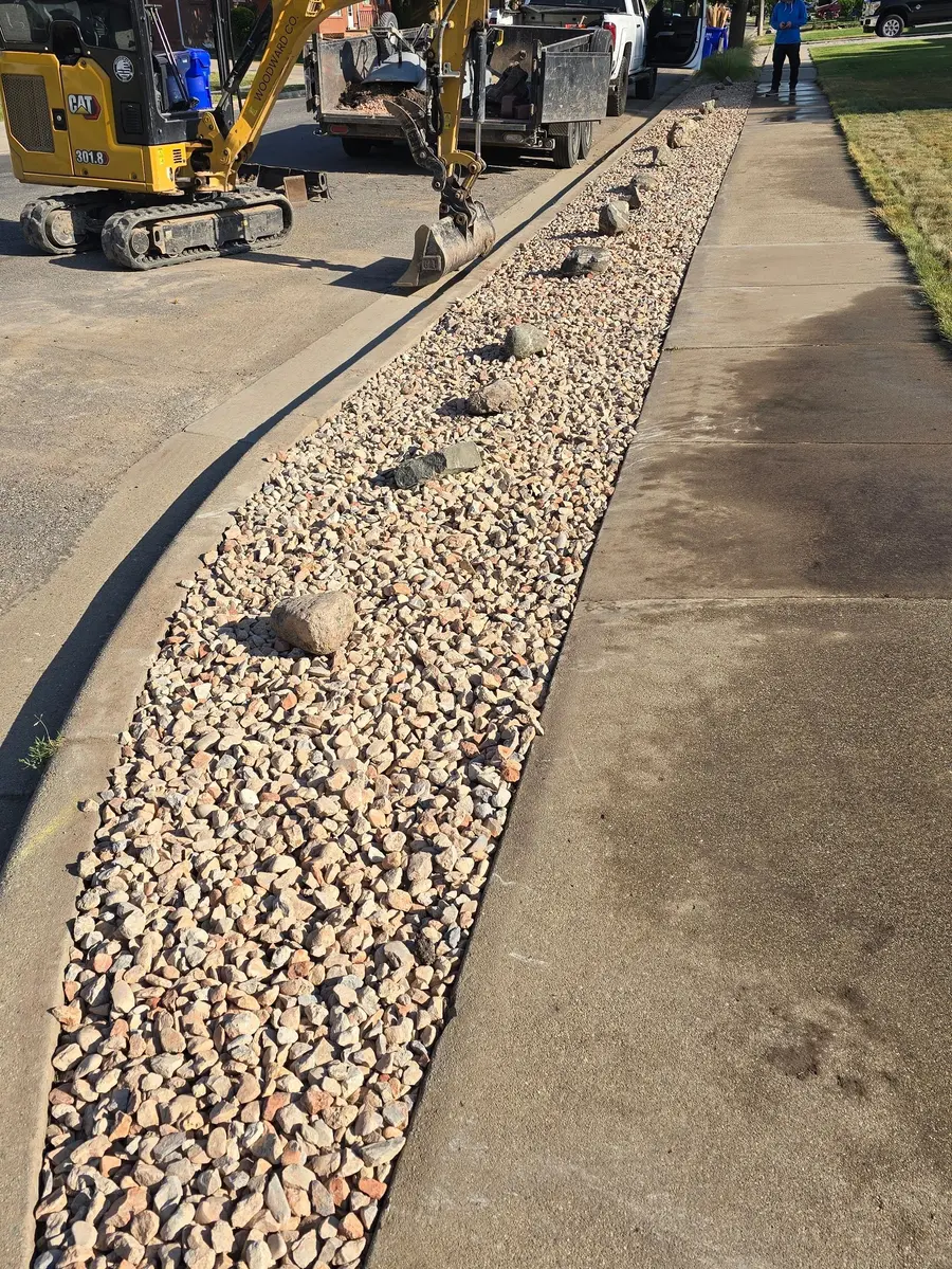 After: the park strip finished in rock mulch with accent boulders, a mini excavator and dump trailer at the curb