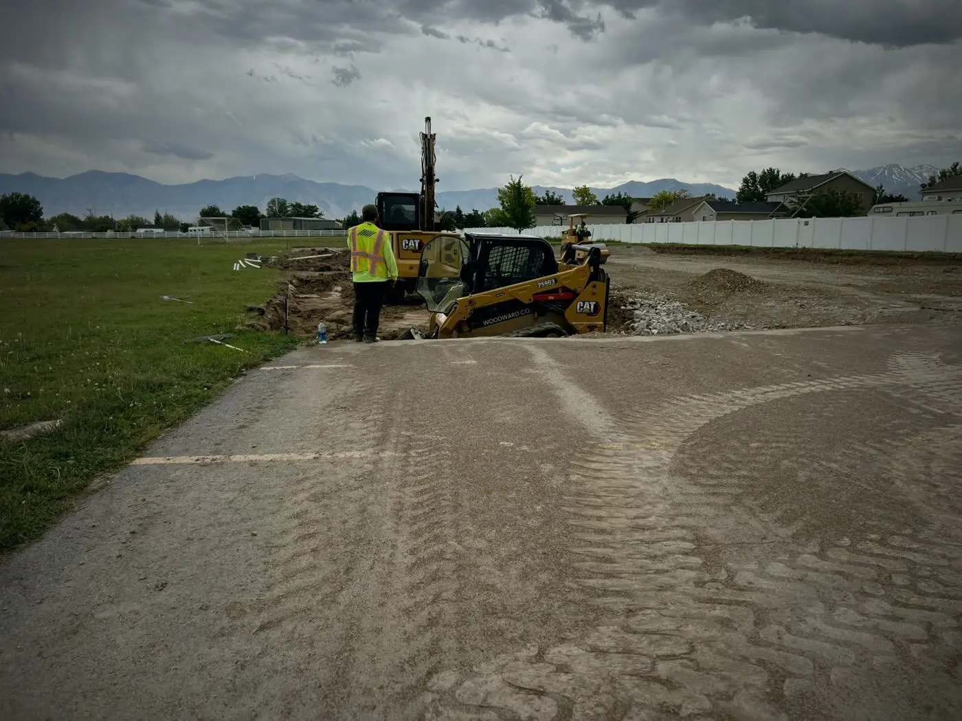 Before: a skid steer and an excavator starting the lot on bare dirt beside an old asphalt path, a crew member in a hi-vis vest, storm clouds over the mountains