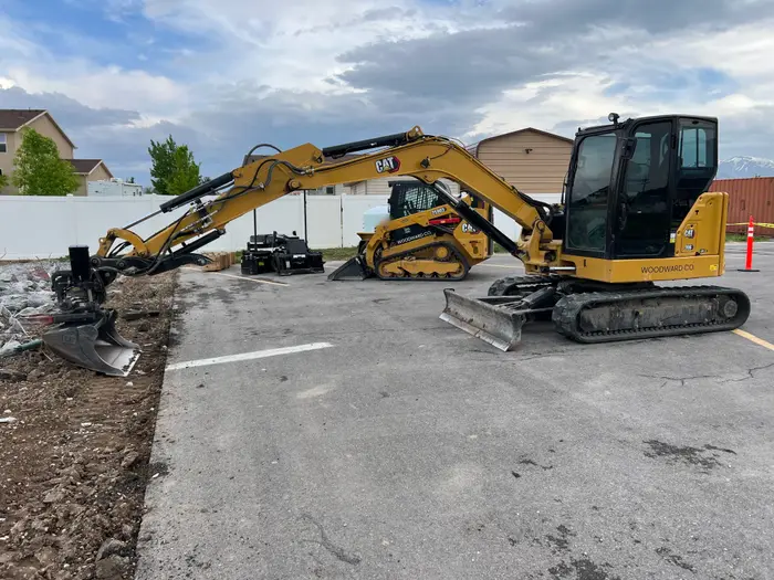 A mini excavator with its arm stretched along the edge of an asphalt lot, a compact track loader and grader box behind