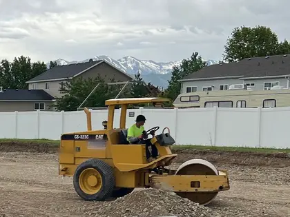 A roller operator compacting road base on a lot, a white vinyl fence and houses behind
