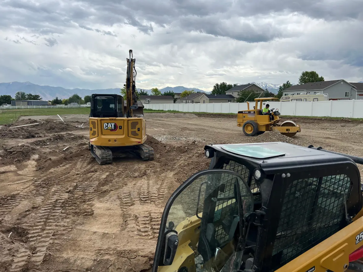From a skid steer cab: a mini excavator and a smooth-drum roller prepping a big dirt lot under storm clouds, houses and mountains behind