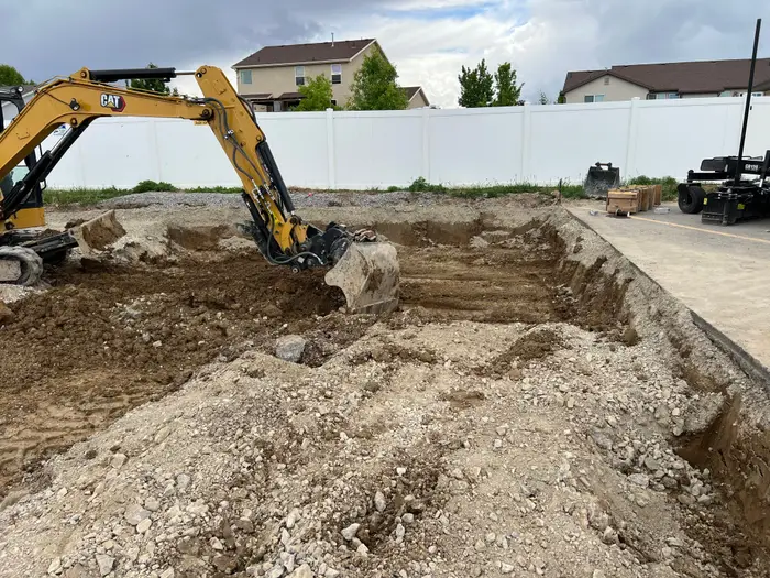A mini excavator digging out subgrade beside existing asphalt, a white vinyl fence and houses behind