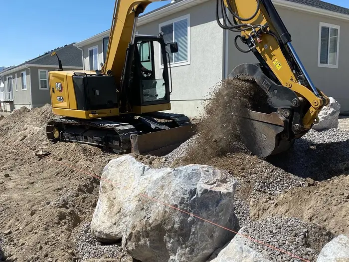 A mini excavator dumping dirt behind a boulder retaining wall between new homes, a string line across the rocks