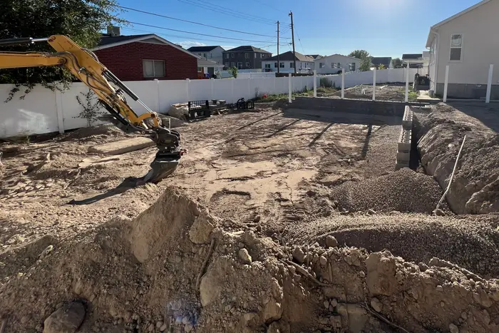 A new-home backyard dug out for a retaining wall, the first blocks along the trench and a mini excavator arm on the left