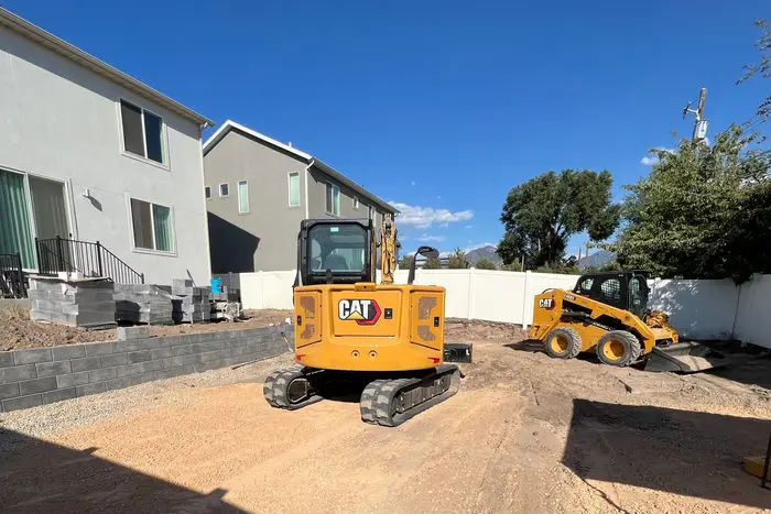 A mini excavator and a skid steer on a graded backyard beside a finished block retaining wall, pallets of block by the house