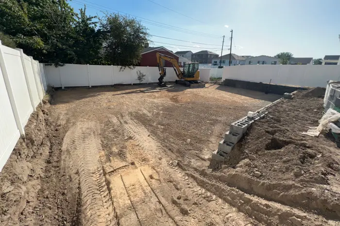 A graded backyard with tire tracks, a mini excavator at the back fence and a cinder block wall going in along the side