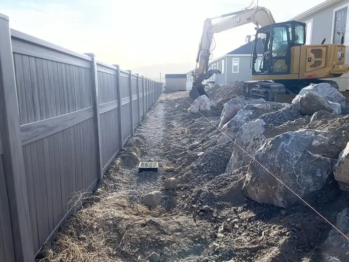 A mini excavator setting boulders for a retaining wall along a vinyl fence in a side yard, a string line and a drain box in the gravel