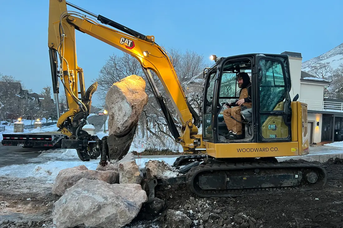 A yellow CAT mini excavator lifting a big boulder with its thumb on a snowy evening, the operator in the cab and homes behind
