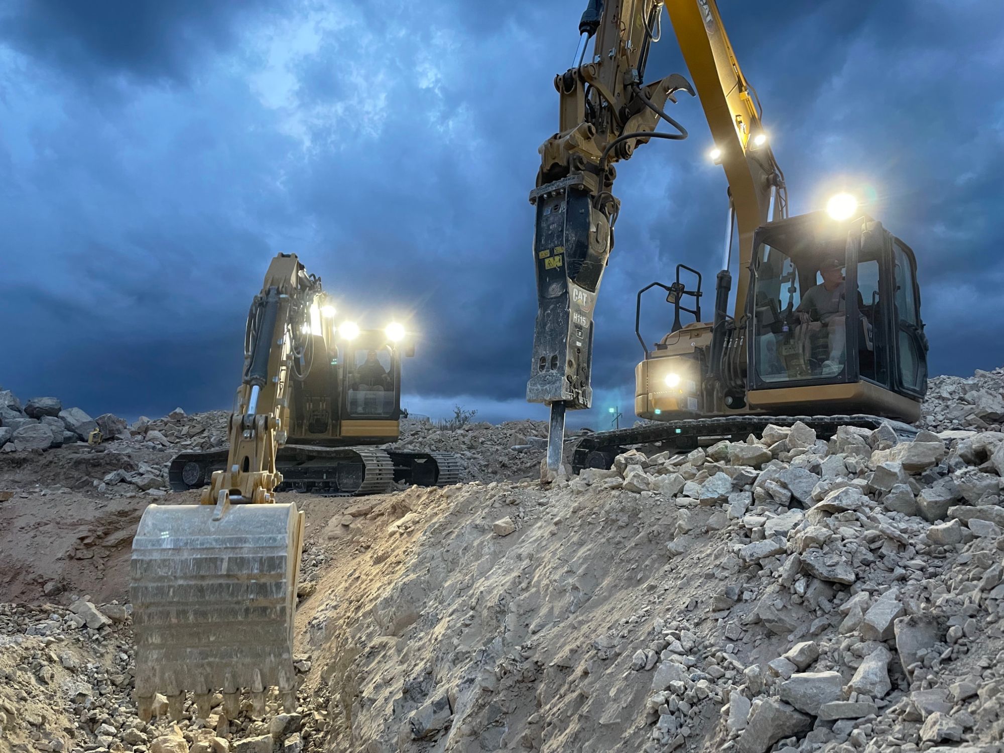 Two excavators working rock under a stormy dusk sky, work lights blazing